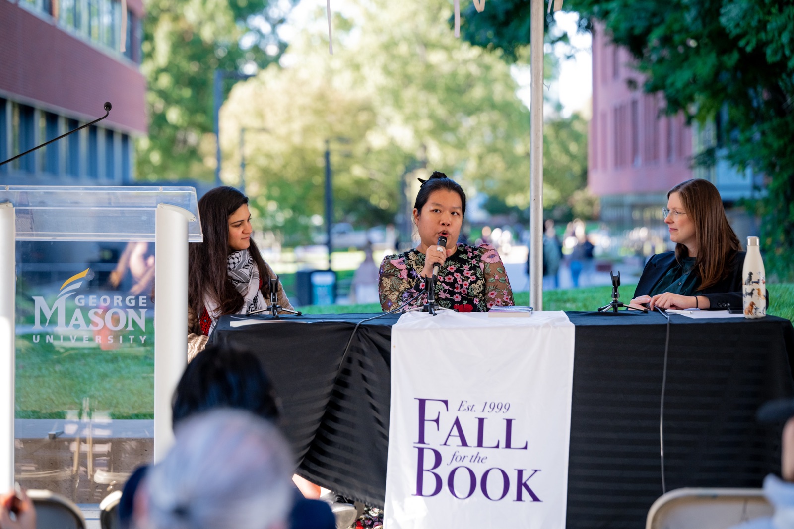 Catherine Olien moderating an outdoor author panel under a tent at George Mason University, listening as a panelist speaks