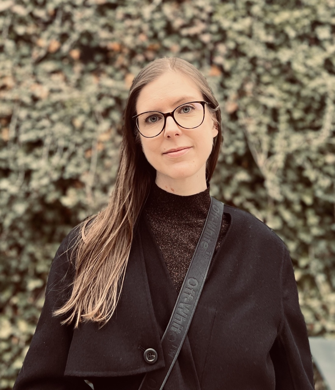 Catherine Olien, in a dark coat and glasses, standing before a wall of ivy