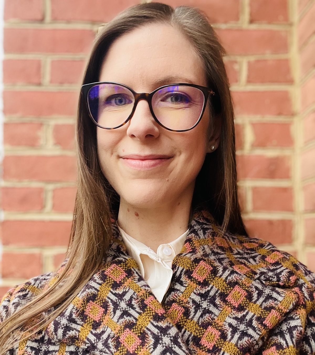 Catherine Olien in glasses and a patterned wool coat, smiling, against a brick wall